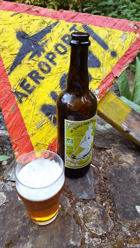 Bouteille et verre de la bière blonde des Faucheurs Volontaires d'OGM dans le jardin devant le panneau "Non à l'aéroport de Notre-Dame des Landes (NDDL)".