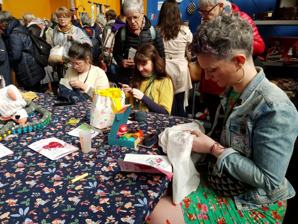 Trois femmes brodent et font du crochet sur le Coin des Blogueuses du salon Pour l'Amour du Fil en 2024. Des visiteuses les regardent avec curiosité.